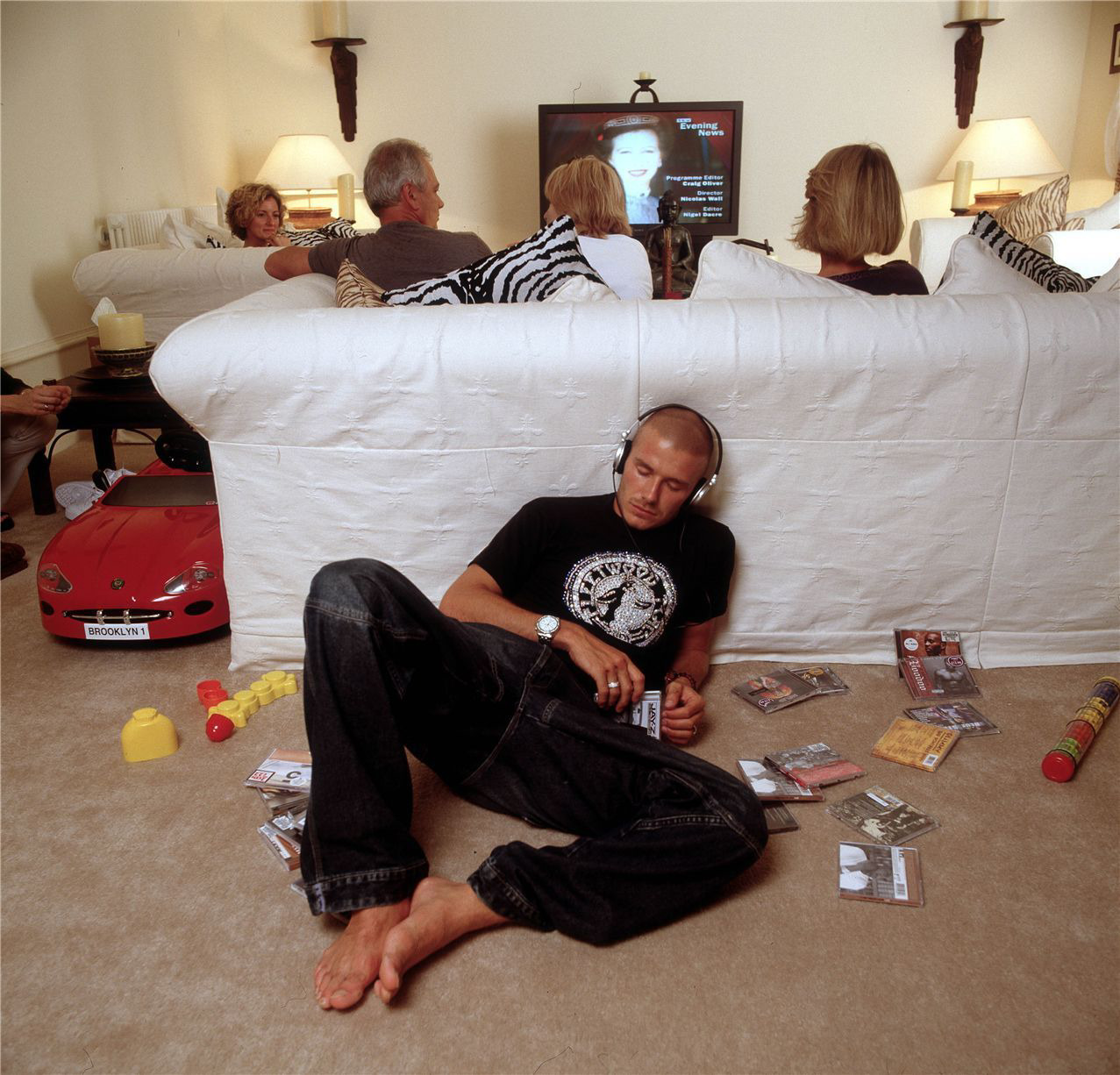 Imagem do jogador David Beckham deitado no chão atrás de um sofá branco, ele é branco, e está com roupas pretas, está com fones de ouvido e de olhos fechados, com diversas capas de discos ao seu redor, ele parece ouvir música