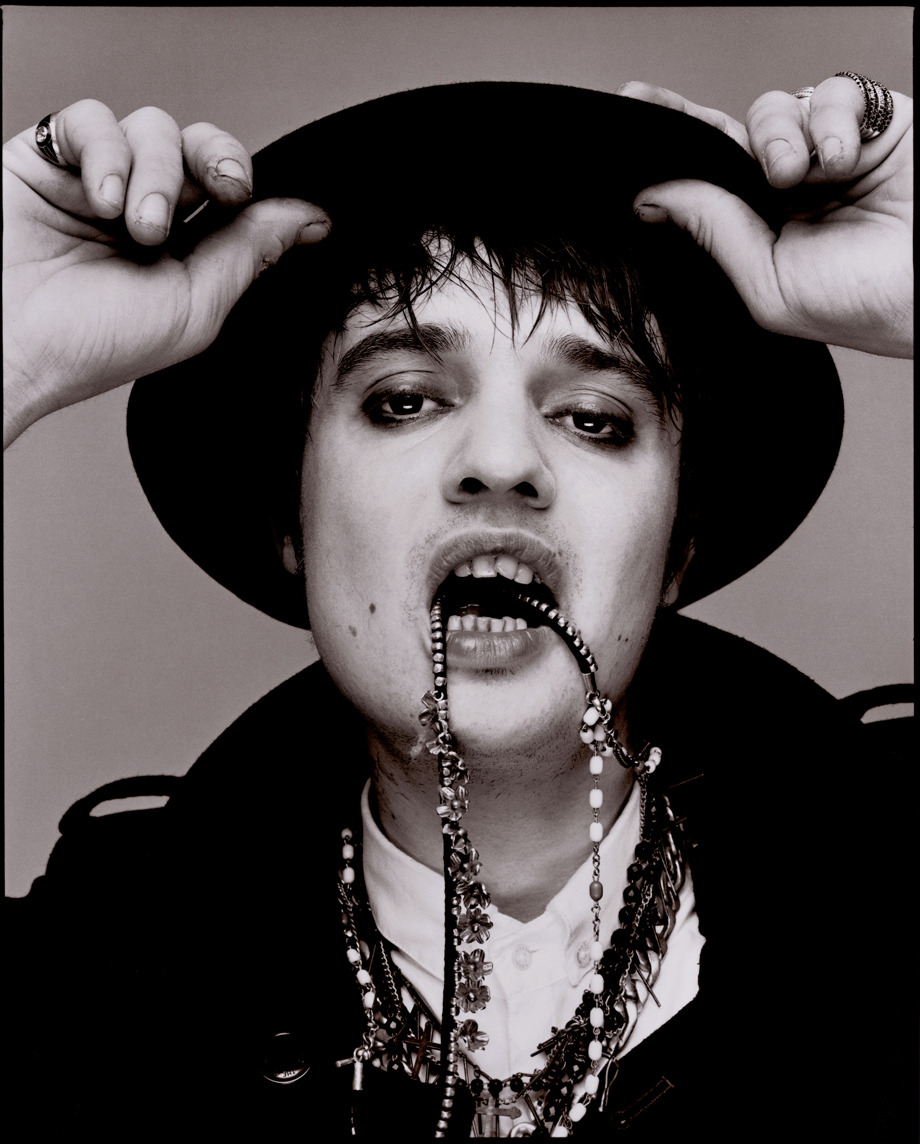 Pete Doherty, homem branco, usando e segurando um chapéu redondo com as duas mãos, ele está com um colar que usa na boca e, veste um blazer preto e camiseta branca, fotografado pela Clash Magazine em 2009
