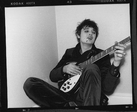 Pete Doherty, sentado com um cigarro na boca, sentado no chão com uma guitarra Rickenbacker