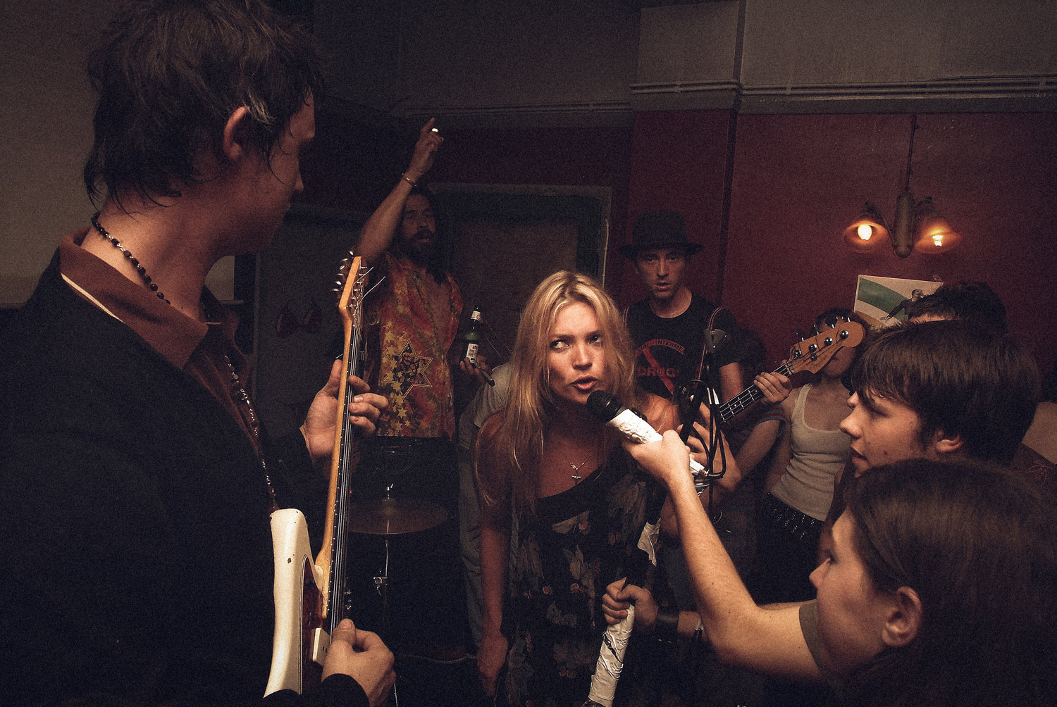 Um lugar com diversas pessoas, entre elas Pete tocando guitarra e sua então namorada na época Kate, mulher branca de cabelos loiros, cantando num microfone segurado por um jovem rapaz branco de cabelo médio castanho escuro.