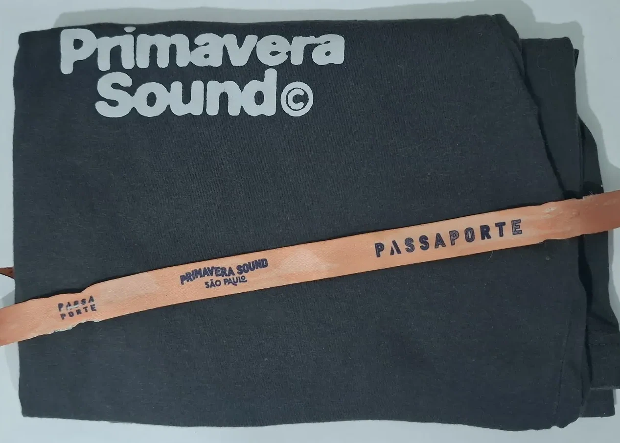 Imagem da camiseta dobrada que comprei no primeiro dia do festival, em cima dela está a pulsera do passaporte