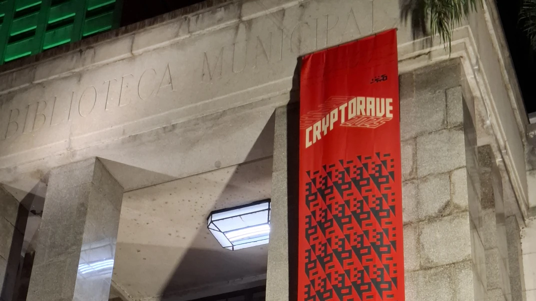 Parte superior da entrada da biblioteca Mario de Andrade, com um banner vermelho longo verticalmente com o logo da edição 2025 da Cryptorave