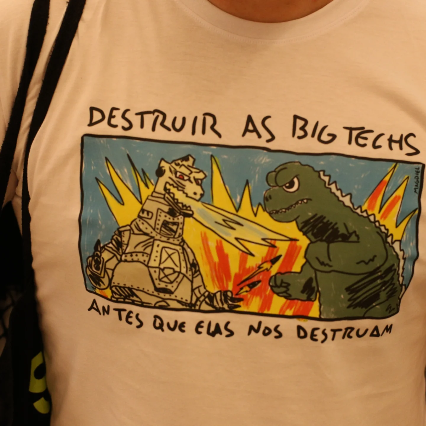 Uma camiseta branco com uma estampa de um godzilla enfretando um robo com a frase &ldquo;destruir as bigtechs antes que elas nos destruam&rdquo;
