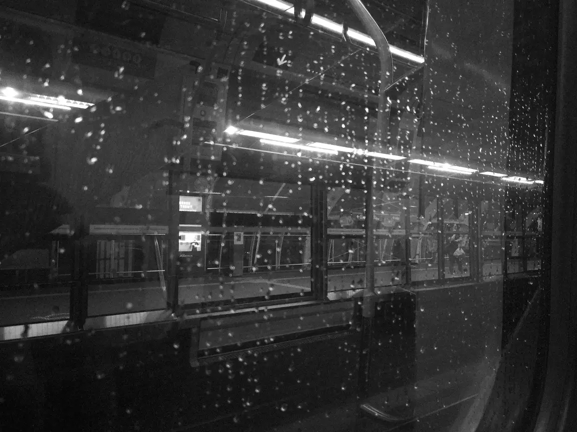 Uma foto em preto e branco da janela do metrô parado na plataforma, com pingos de água da chuva