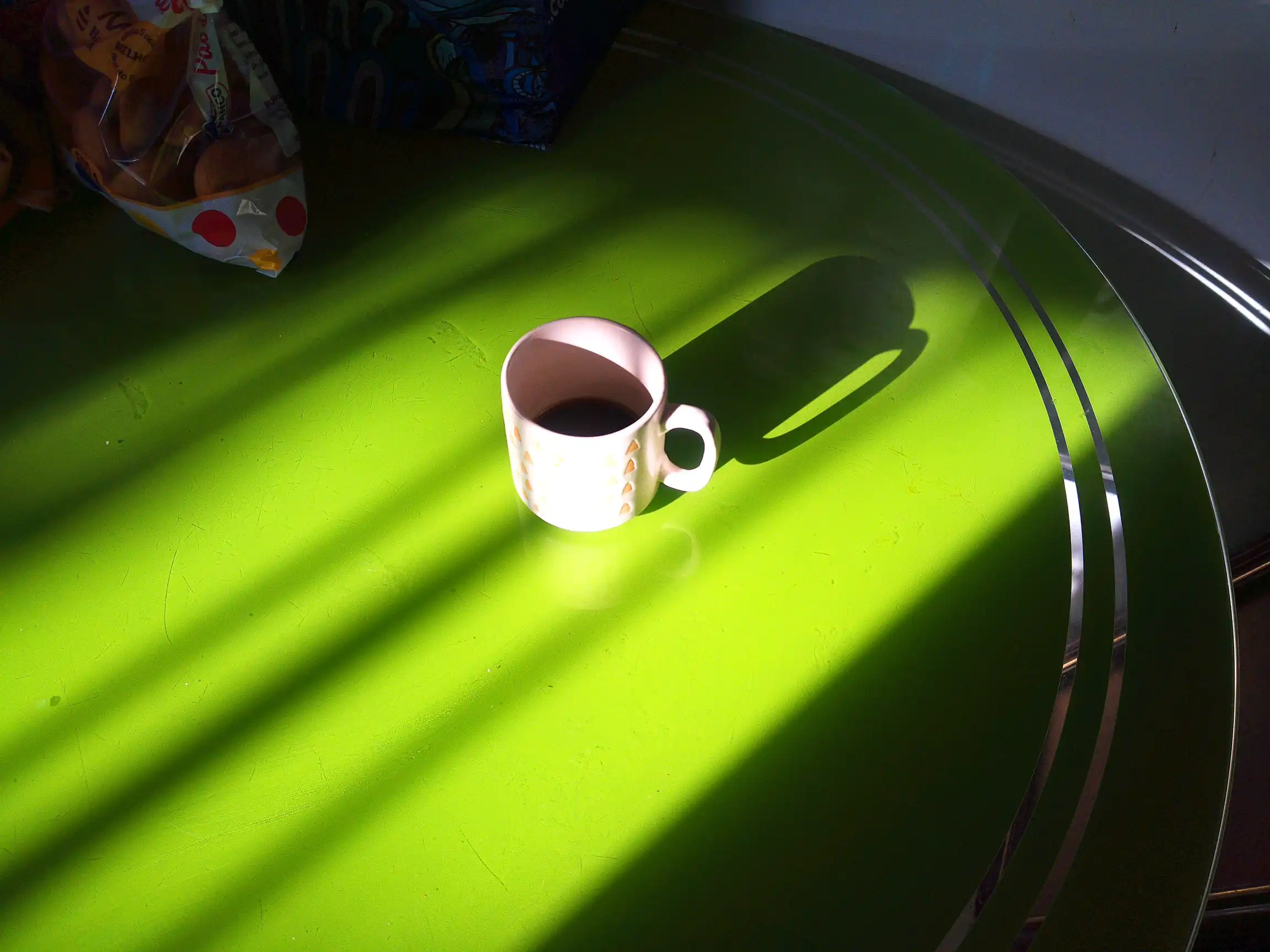 Uma xicará de café, sob uma mesa redonda verde, ilumidada por um feixe luz de uma manhã de sábado