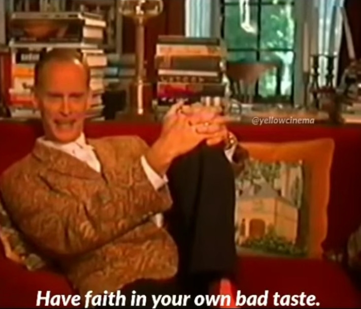 John Waters sentado em um sofá vermelho com a legenda em inglês &ldquo;Tenha fé no seu próprio mal gosto&rdquo;